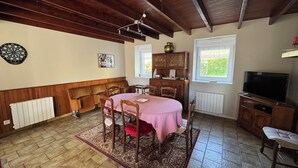 A dining area with a round table covered in a checkered tablecloth, surrounded by wooden chairs. There is a radiator, a dartboard, and a window with a view of greenery.