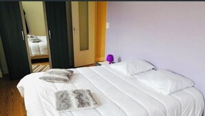 A neatly made bed with white bedding and two pillows, a wooden floor, and a purple lamp on a nightstand.