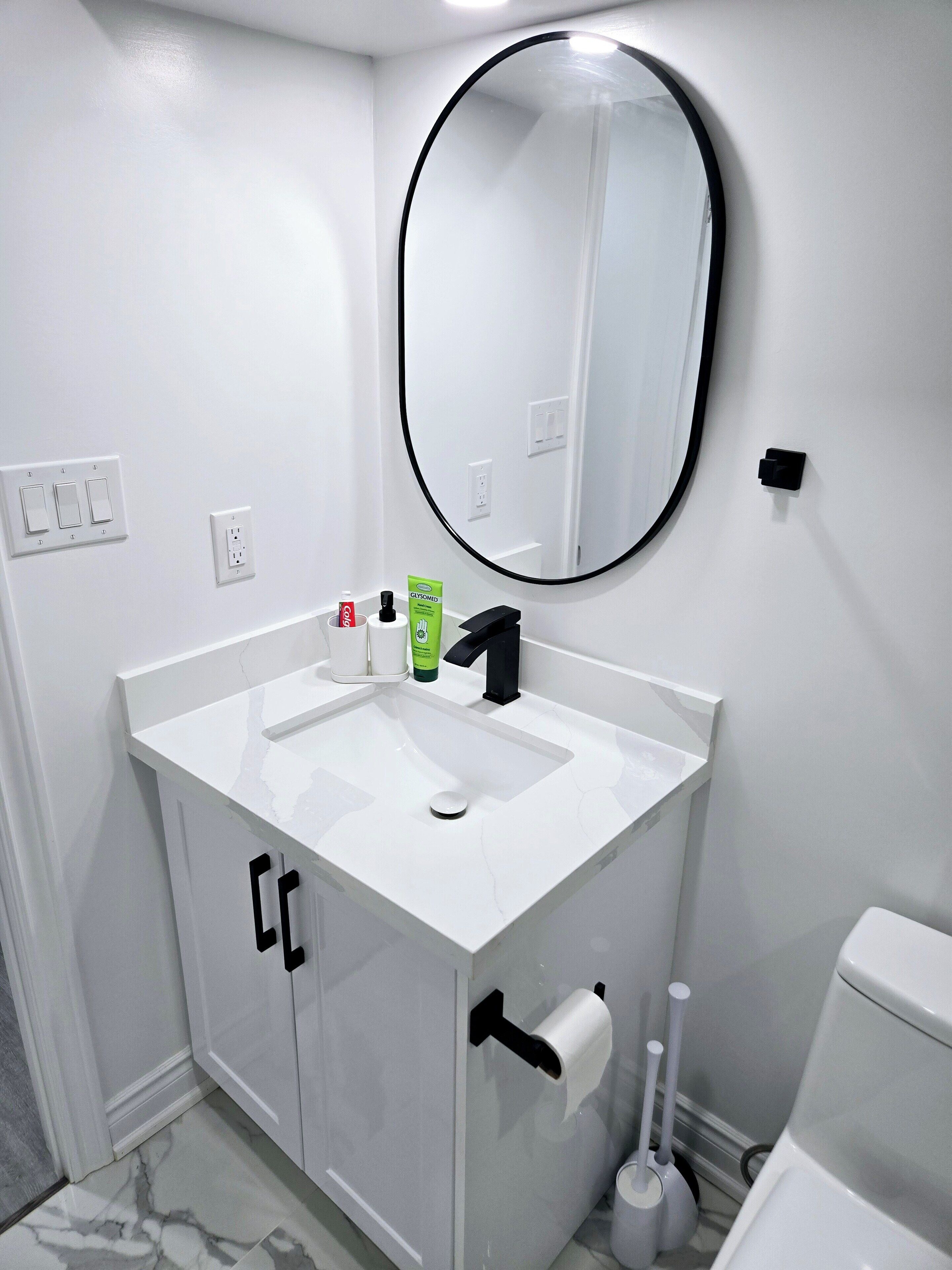 Une salle de bain avec un lavabo blanc, un robinet noir et un miroir rond. On peut voir des articles de toilette sur le lavabo et des toilettes sur le côté droit.