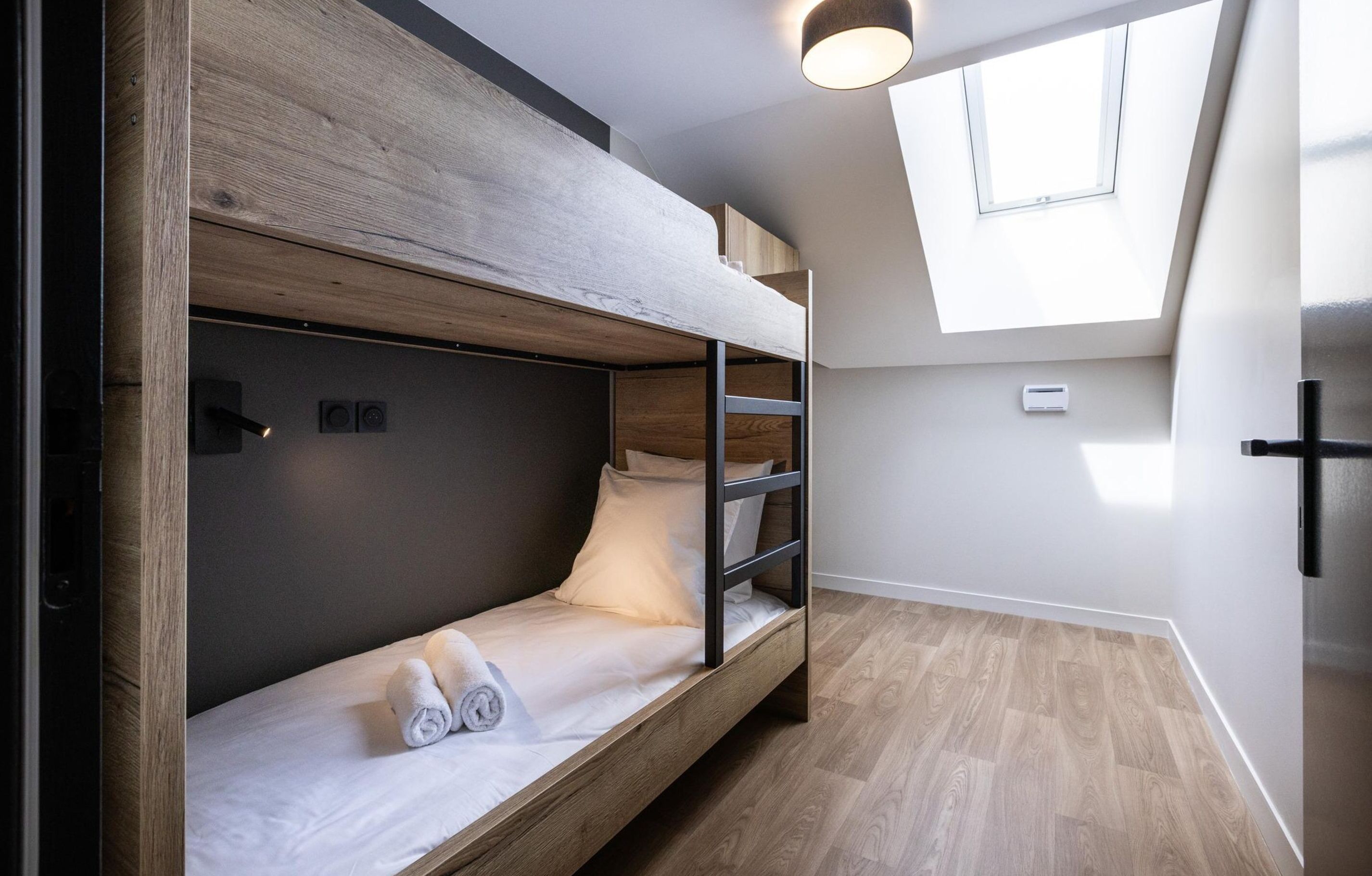 A bunk bed with a skylight above, a small air conditioning unit on the wall, and a wooden floor.