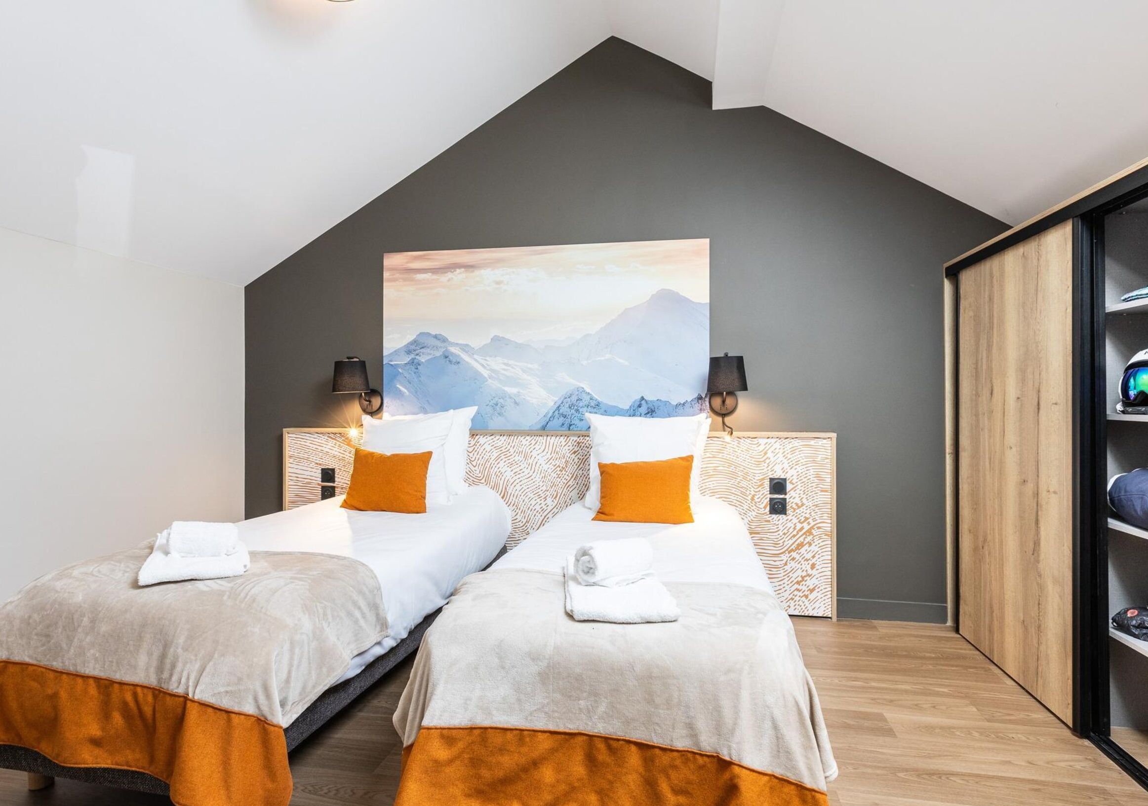 A modern hotel room with two beds, a large mountain painting on the wall, and a wooden wardrobe.
