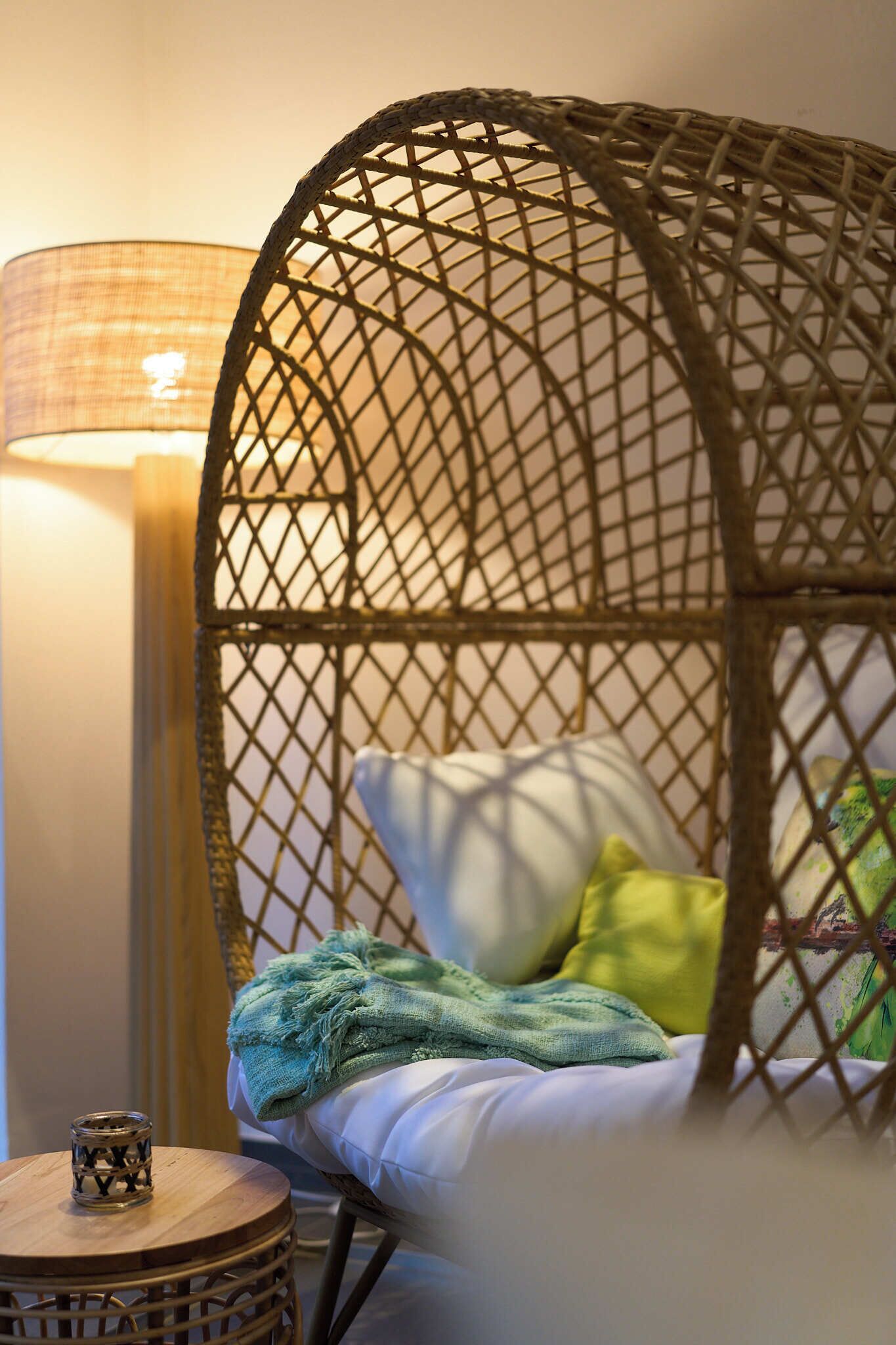 Ein gemütliches Zimmer mit einem Korbstuhl, einem Nachttisch mit Kerze und einer Lampe.