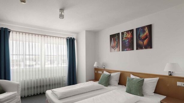 Camera d'albergo con due letti, una finestra con tende leggere, una testiera in legno e tre quadri appesi al muro.