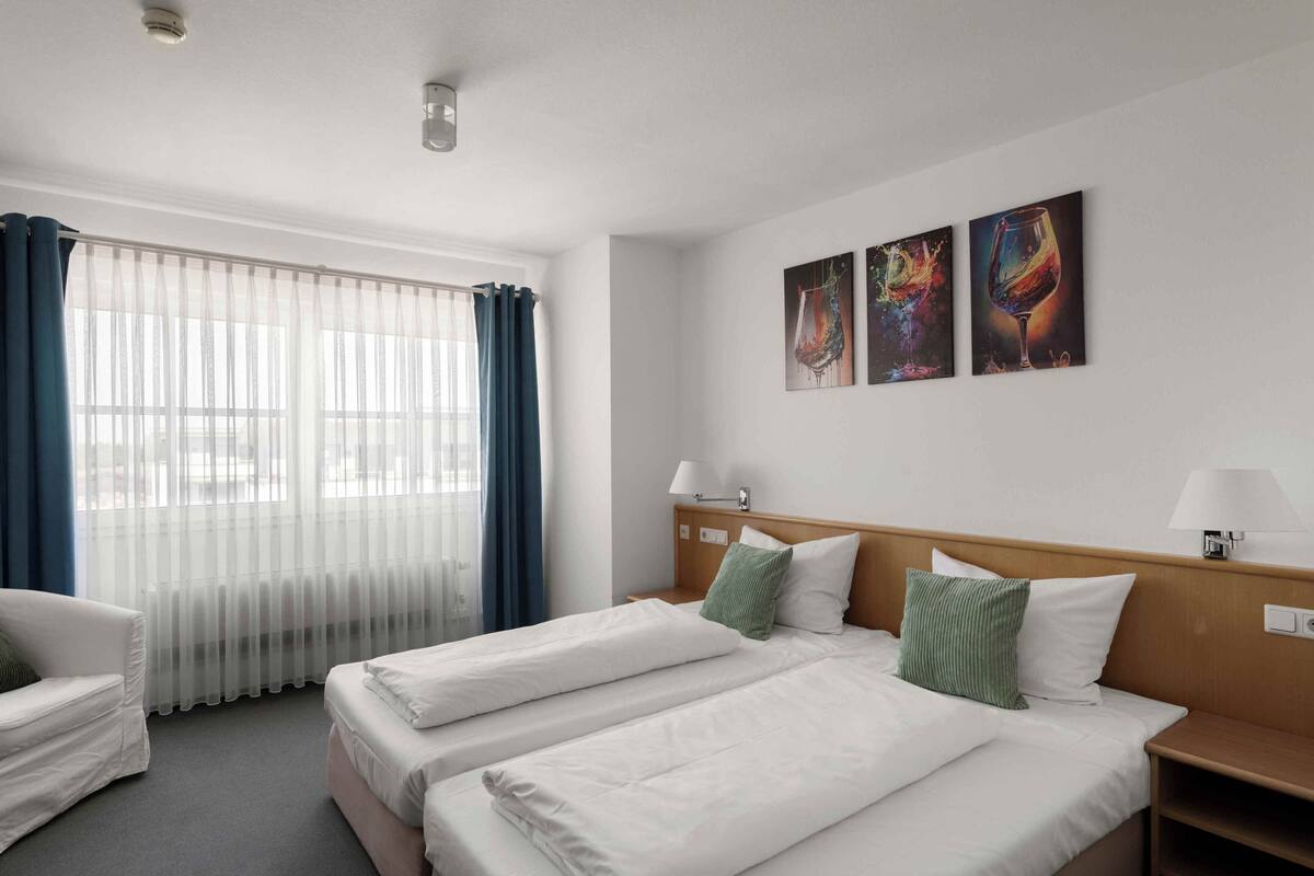 A hotel room with two beds, a window with sheer curtains, a wooden headboard, and three wall-mounted paintings.