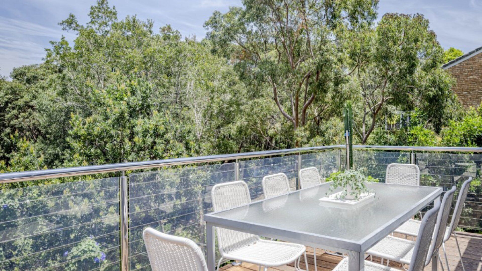 A glass-top table with white wicker chairs on a balcony with a metal railing, surrounded by dense green foliage.