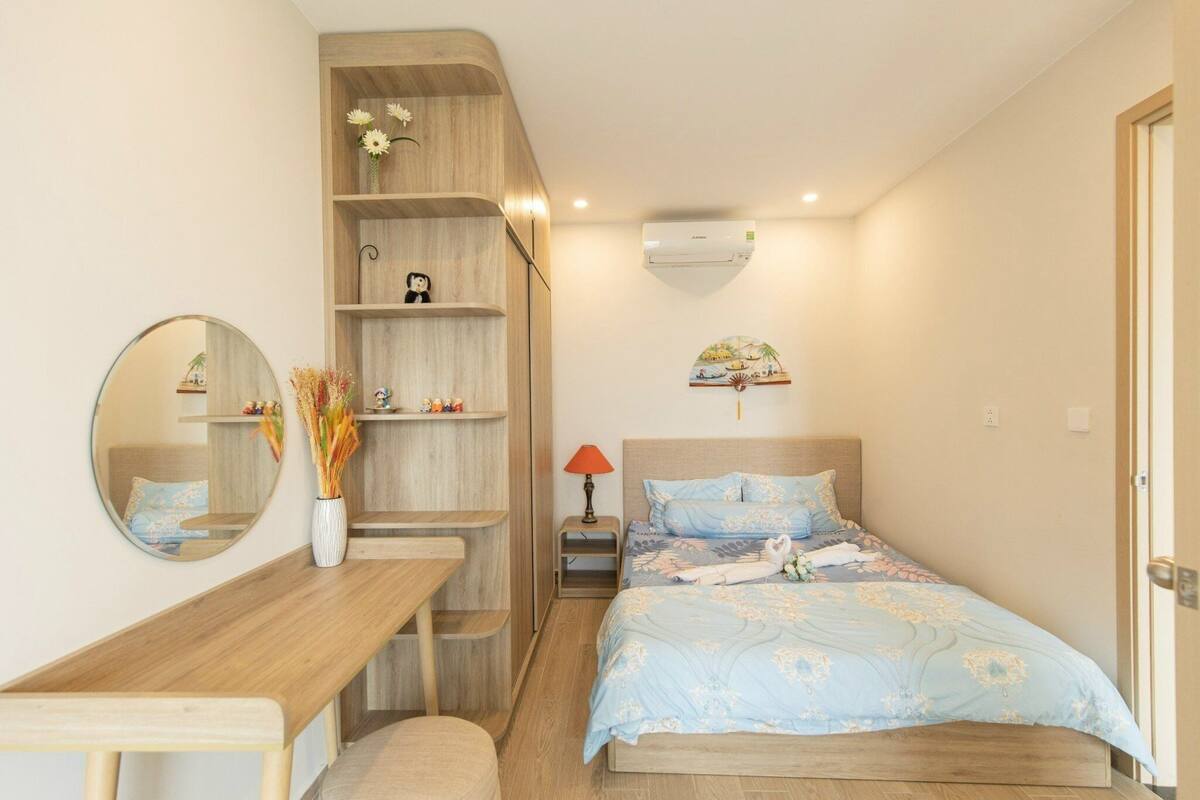 Une chambre avec un bureau en bois, un lit recouvert d’une courtepointe à motifs floraux bleus, un miroir, une étagère avec des objets décoratifs et un climatiseur fixé au mur.