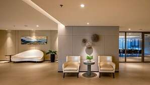 Um lobby moderno com uma cadeira branca curvada, dois poltronas de couro, uma mesa redonda com uma planta e uma grande pintura abstrata na parede.