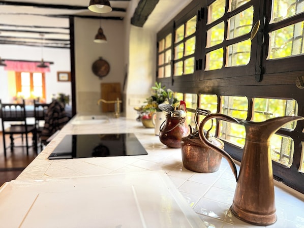 Un plan de travail de cuisine avec des théières en cuivre et une bouilloire, une fenêtre donnant sur un paysage verdoyant, et un coin repas à l’arrière-plan.