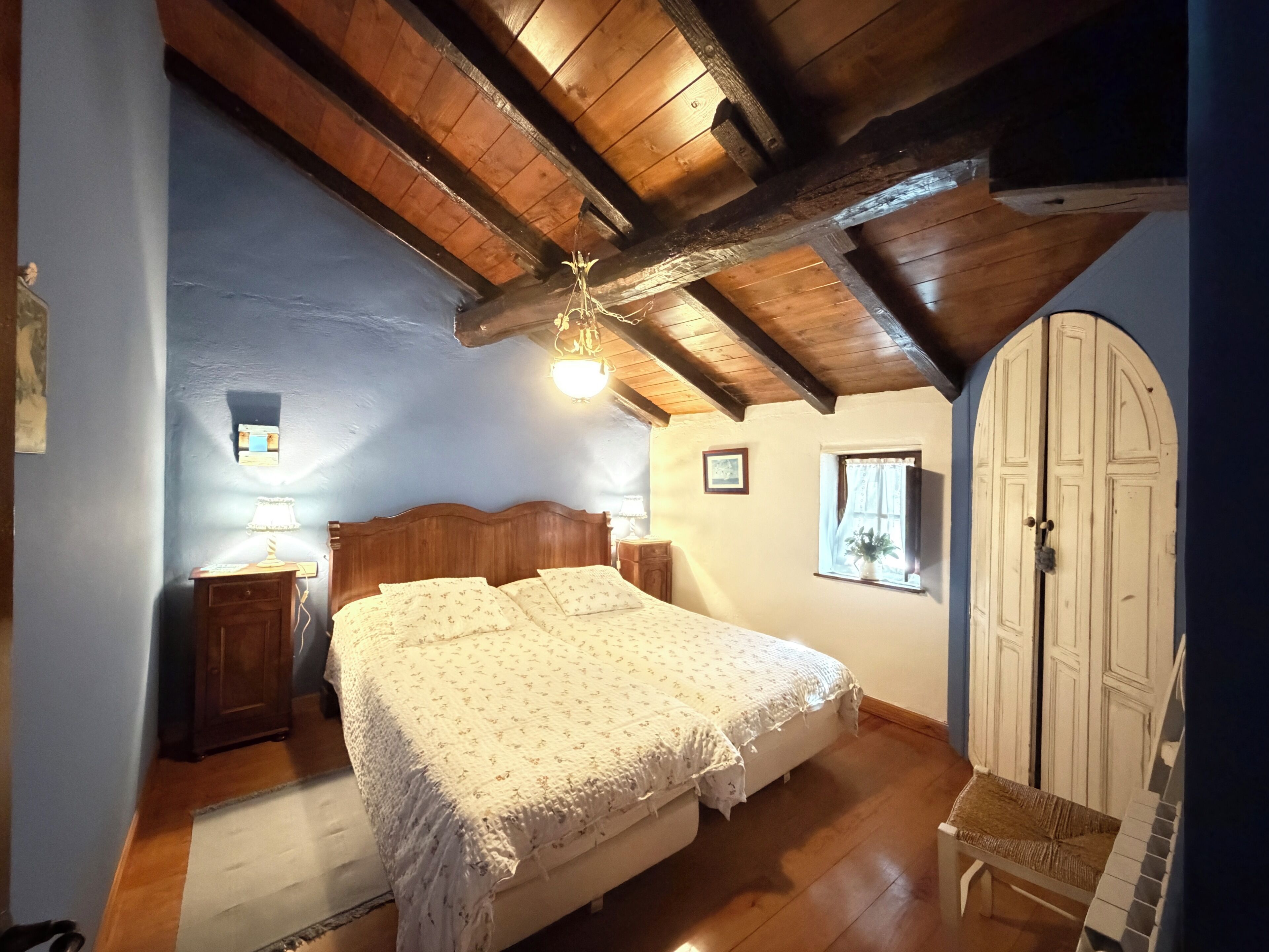 A bedroom with a wooden ceiling, a bed with a floral bedspread, a nightstand, a window with curtains, and a door.