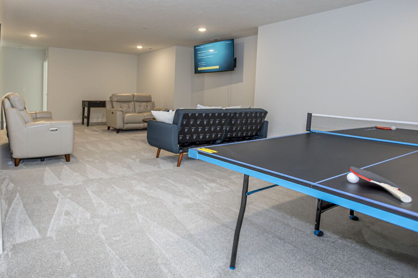 A ping pong table in a room with a sofa, a chair, and a TV mounted on the wall.