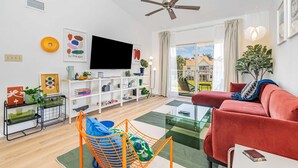 Un salon moderne avec un canapé rouge, une table basse en verre et un ventilateur de plafond. Un téléviseur est fixé au mur et une grande fenêtre offre une vue sur l’extérieur.