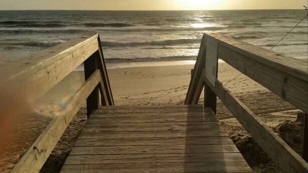 Una pasarela de madera que lleva a una playa con el sol poniéndose al fondo.