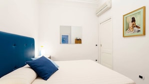 Un dormitorio con cama, cabecera azul, pared blanca, una puerta, unidad de aire acondicionado, un cuadro enmarcado y una mesita de noche con lámpara.
