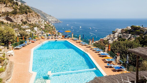 Zona de piscina con sillones de descanso y sombrillas, con vistas a un pueblo costero y al mar.