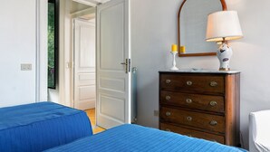 Een slaapkamer met twee blauwe bedden, een houten dressoir met spiegel en lamp, en een witte deur die naar een andere kamer leidt.