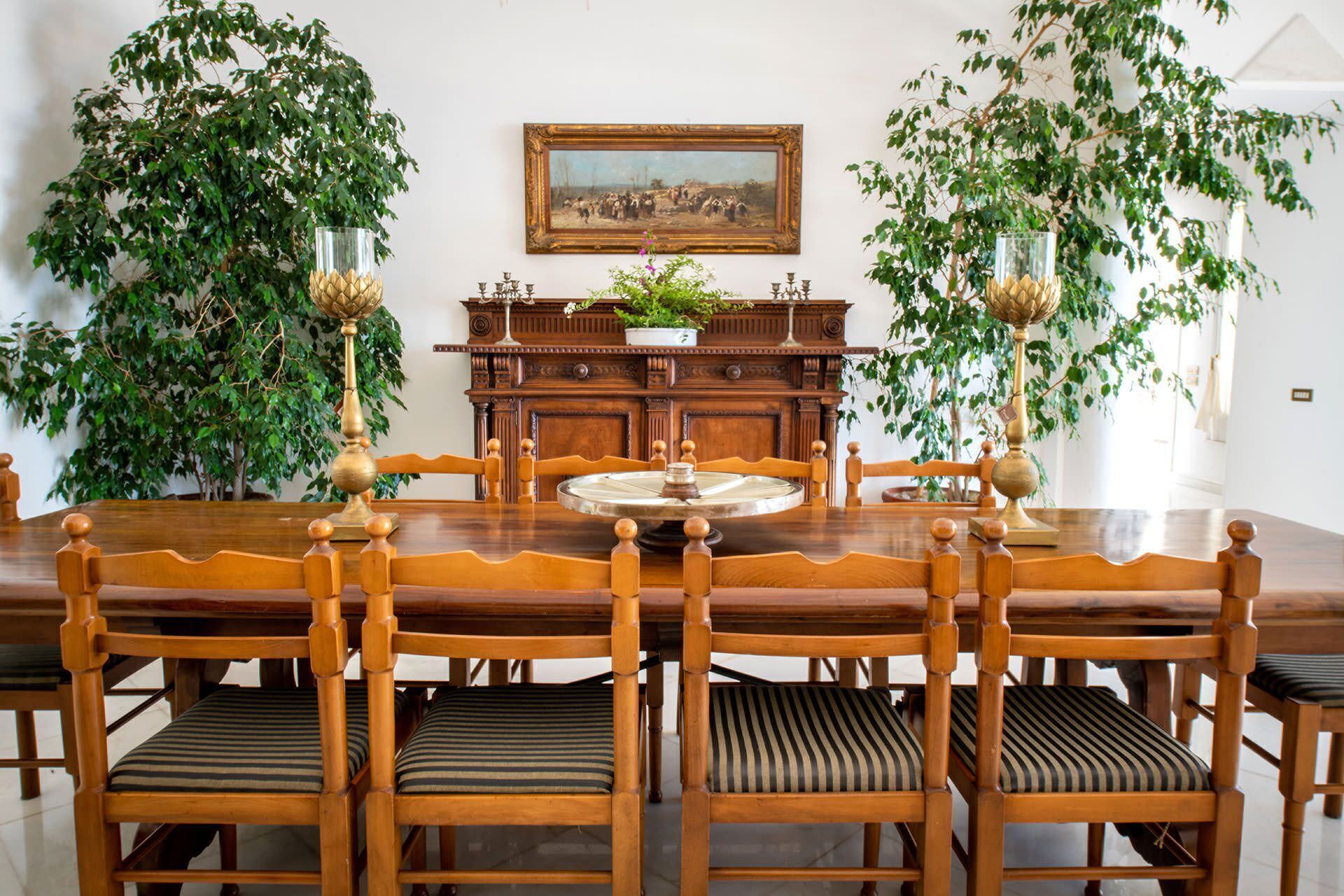 A dining room with a wooden table, six chairs, a cabinet, and two large potted plants.
