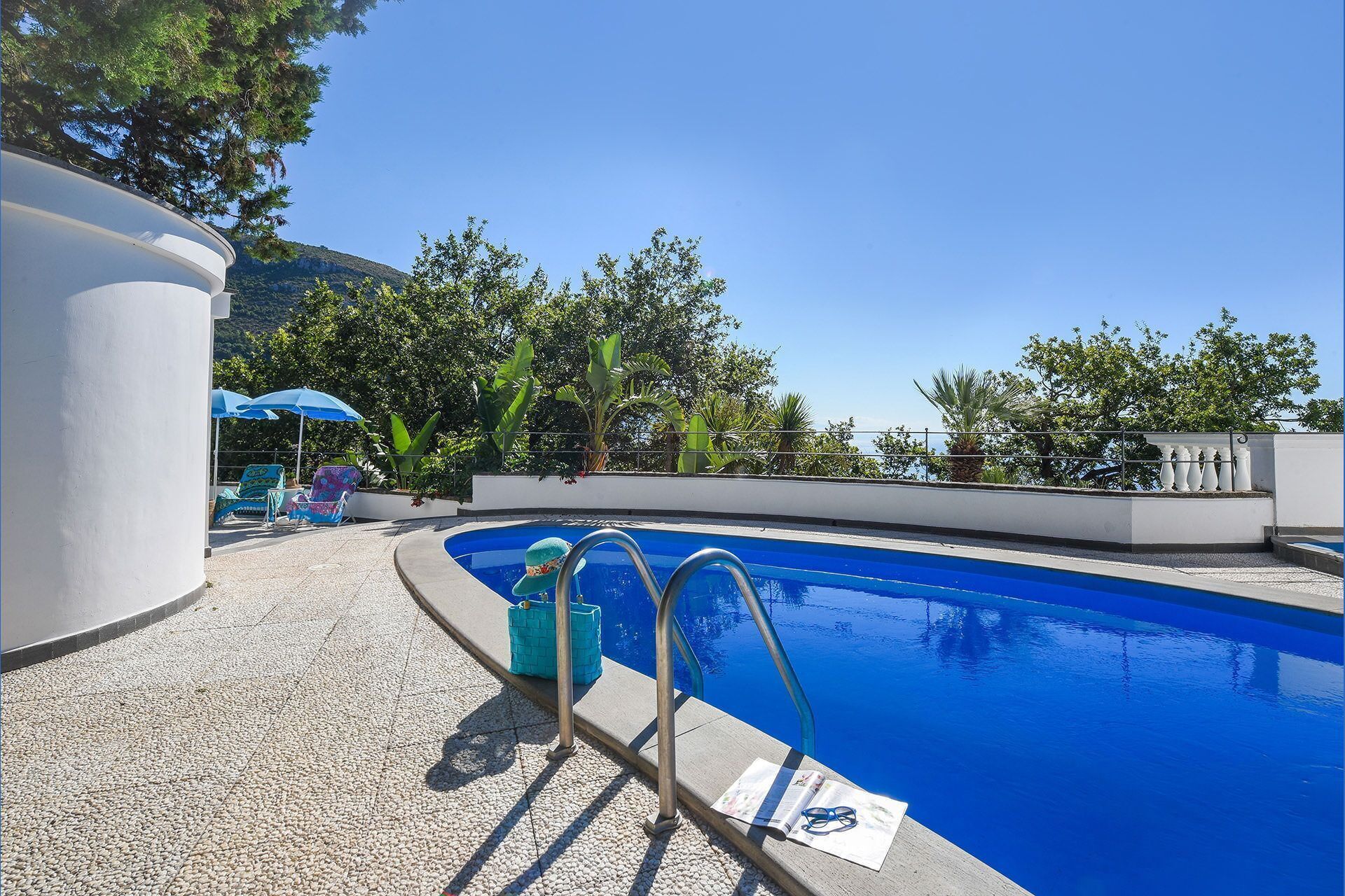 A swimming pool with a clear blue water, surrounded by a white wall and a stone floor. There are lounge chairs and umbrellas in the background.