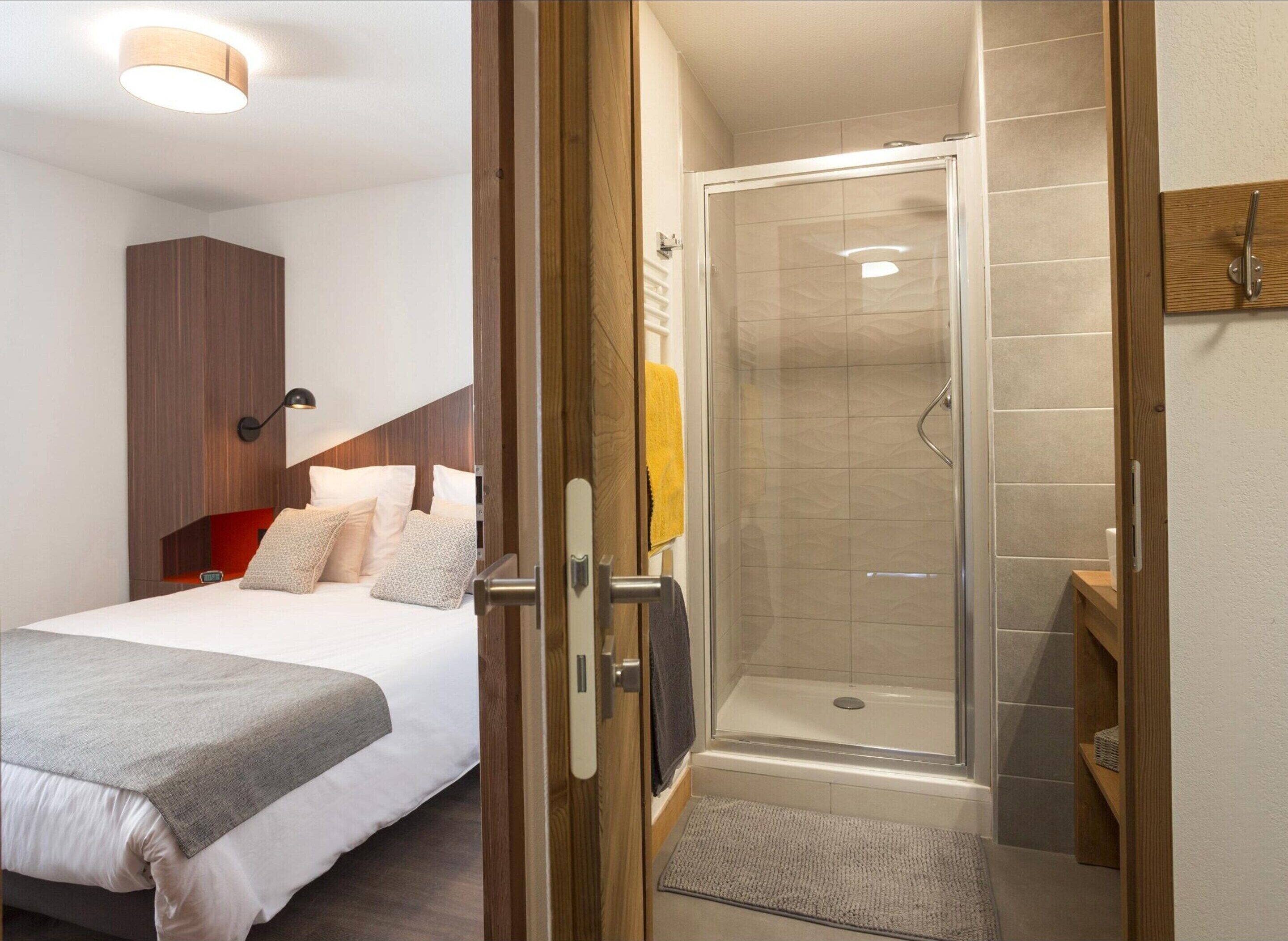 Habitación de hotel con cama, armario de madera, zona de ducha con puerta de cristal y un pequeño estante de madera.