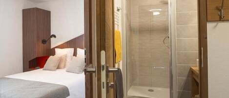 Habitación de hotel con cama, armario de madera, zona de ducha con puerta de cristal y un pequeño estante de madera.
