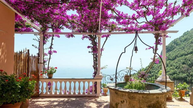 Ein überdachter Außenbereich mit Meerblick, ausgestattet mit einer Pergola, blühenden Pflanzen, einer Fontäne und Kübelpflanzen.