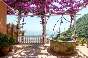 Un espacio al aire libre cubierto con vista al mar, que cuenta con una pérgola adornada con plantas florecientes, una fuente y plantas en macetas.