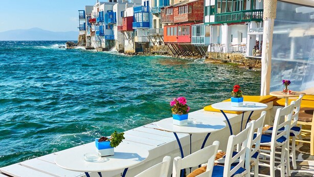 A seaside restaurant with outdoor seating overlooking the sea, featuring white tables and chairs, and colorful buildings in the background. - Rose Studio Two with shared pool and sea view (Mykonos)