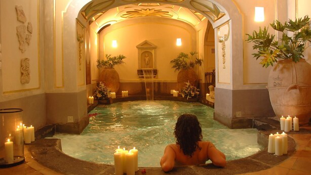 Una persona relajándose en un jacuzzi rodeado de velas y plantas, con una estatua y un techo decorado al fondo.