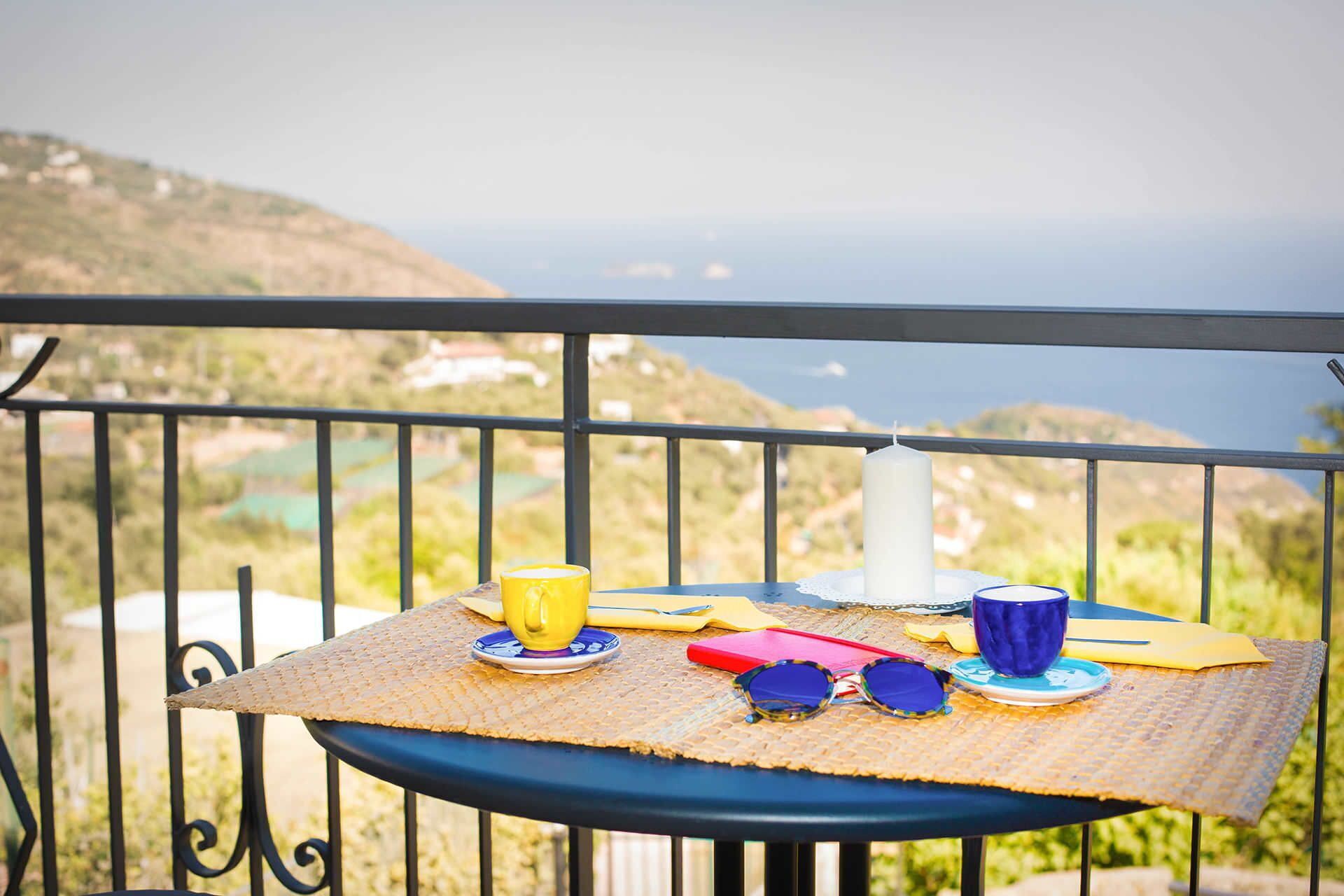 Ein Tisch mit einer Kerze, einer gelben Tasse, einer blauen Tasse und einer Sonnenbrille auf einem Balkon mit schöner Aussicht.