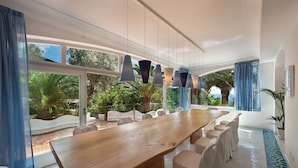 A spacious dining area with a long wooden table and white chairs, large windows with blue curtains, and hanging pendant lights. - Villa delle Sirene with pool - My Rental Homes (Massa Lubrense)