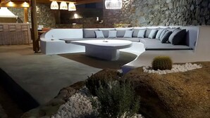 A modern outdoor seating area with a curved sofa and a central coffee table, surrounded by a stone wall and a small cactus plant. - My Rental Homes - Villa Pirgi with swimming pool and panoramic sea views. (Paraga)