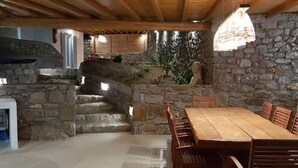 A rustic dining area with a stone wall, wooden staircase, and a table with chairs. - My Rental Homes - Villa Pirgi with swimming pool and panoramic sea views. (Paraga)
