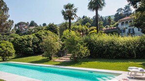 Una piscina con agua azul cristalina, rodeada de abundante vegetación y palmeras. Hay sillas de descanso y una pequeña mesa con un sombrero y una hogaza de pan.