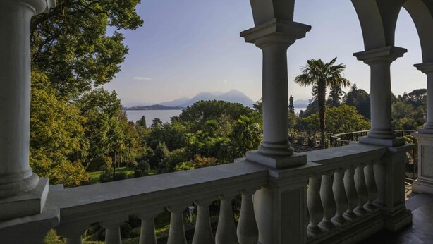 Un balcón de arquitectura clásica con vistas a un jardín exuberante y una cadena montañosa distante.