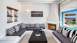 A modern living room with a sectional sofa, a coffee table, and a fireplace. There is a large window with a view of the sea and a beach. - My Rental Homes - Villa Tramountana with pool and large outdoor spaces with BBQ (Fira)