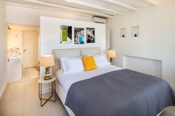 Une chambre d’hôtel moderne avec un grand lit, deux lampes de chevet, une tête de lit ornée d’une œuvre d’art, une petite table avec une lampe et une salle de bain équipée d’un lavabo et d’un porte-serviettes.