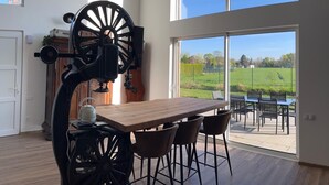 Un coin repas avec une grande table et des chaises, un ventilateur d’époque et une vue sur un champ verdoyant grâce à de grandes fenêtres.