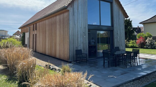 Une maison moderne dotée d’une grande baie vitrée, d’un patio avec mobilier d’extérieur et d’une façade en bois.