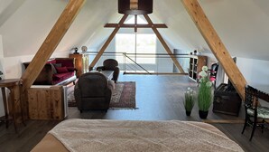 Une pièce mansardée spacieuse avec un plafond en pente, des poutres apparentes, une grande fenêtre, un coin salon confortable avec un canapé et des fauteuils, et une petite table.