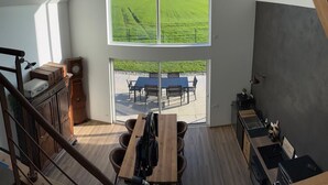 Un espace de vie de style « loft », doté d’une grande fenêtre donnant sur un champ verdoyant, d’un coin repas avec une table et des chaises, et d’un escalier avec une rampe en bois.
