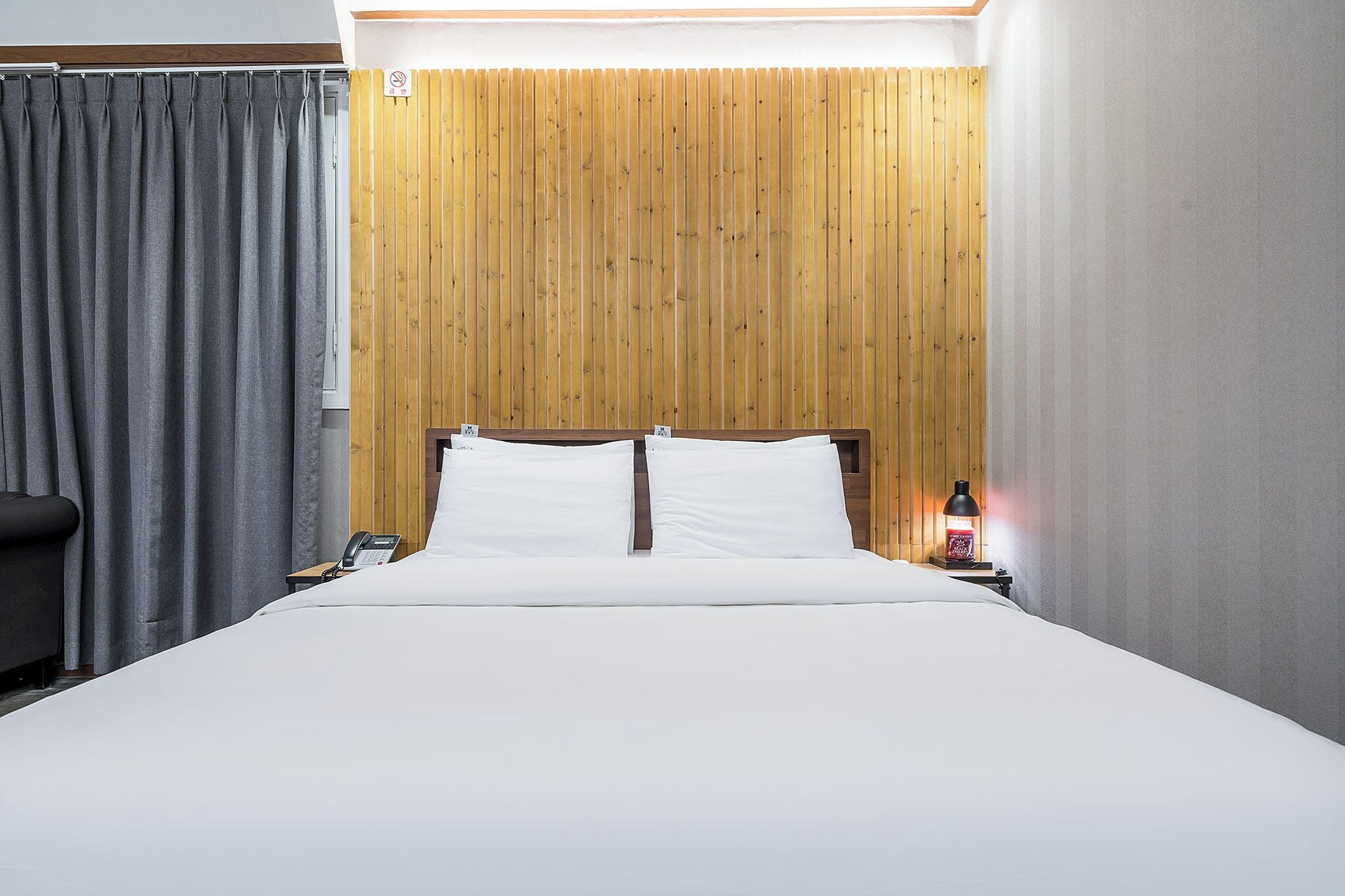 A hotel room with a bed, a wooden headboard, a nightstand with a lamp, and a grey curtain.