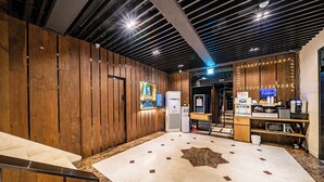 A modern hotel lobby with wooden paneling, a reception desk, a coffee machine, and a digital display. - SKYHOTEL (Yongin)
