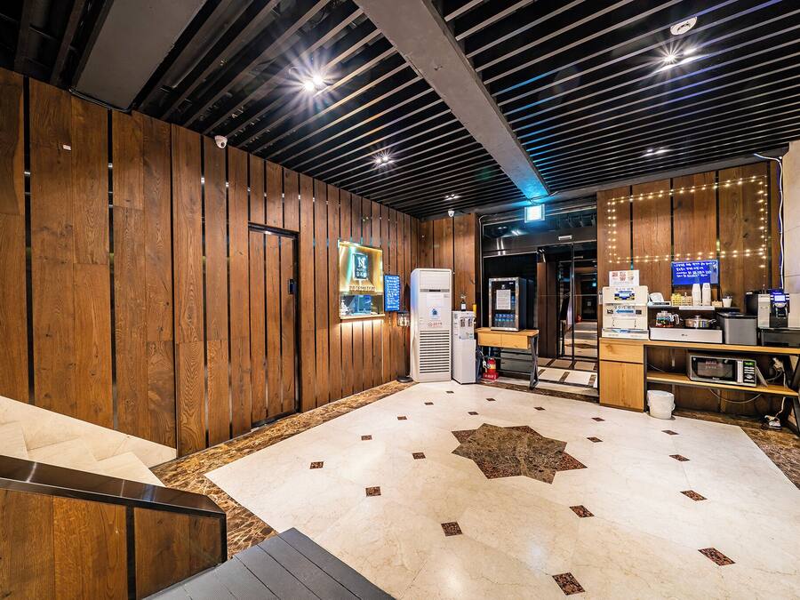 A modern hotel lobby with wooden paneling, a reception desk, a coffee machine, and a digital display.