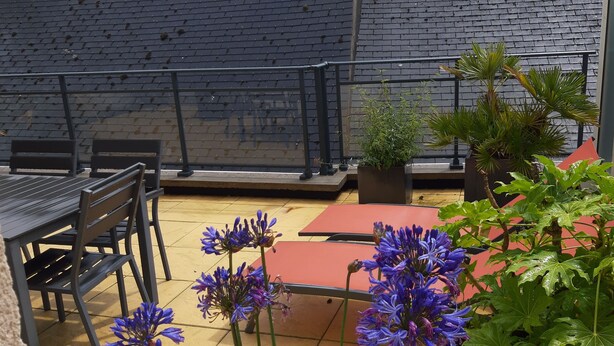 Un área de estar al aire libre con una mesa y sillas de metal negro, rodeada de macetas con plantas y una mesa roja.