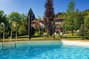 A swimming pool with a wooden deck and metal ladder, surrounded by lush greenery and a house with a red roof.