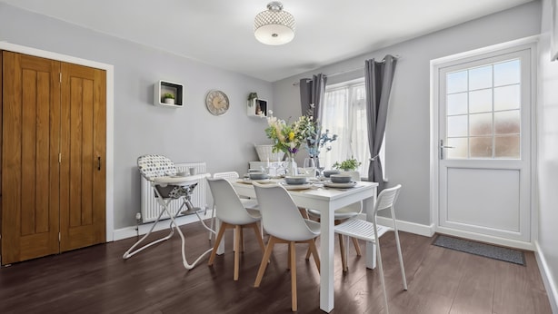 A dining area with a white table set for four, surrounded by white chairs with wooden legs. A white radiator is visible to the left, and a wooden door is on the left side of the room.