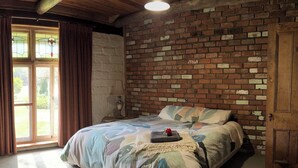 Ein Schlafzimmer mit einer Ziegelwand, einem Bett mit blumigem Überwurf, einer Holztür und einem Fenster mit Vorhängen.