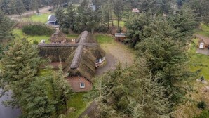 Une maison à toit de chaume, dont le toit a la forme d’une croix, entourée d’une forêt dense et d’un petit étang.