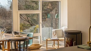 Un coin repas avec une table en bois, des chaises et une vue sur les arbres à travers de grandes fenêtres.