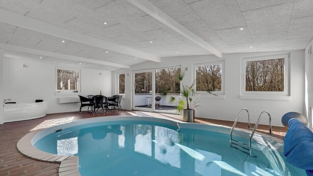 An indoor swimming pool with a clear view of the outside, a seating area, and a window with a view of trees. - Holiday home with pool and spa close to the Limfjord. (Roslev)