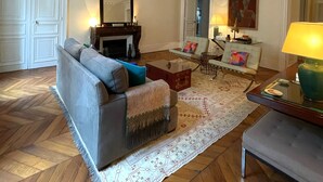 A living room with a sofa, armchair, and a coffee table. There is a fireplace, a mirror, and a painting on the wall.